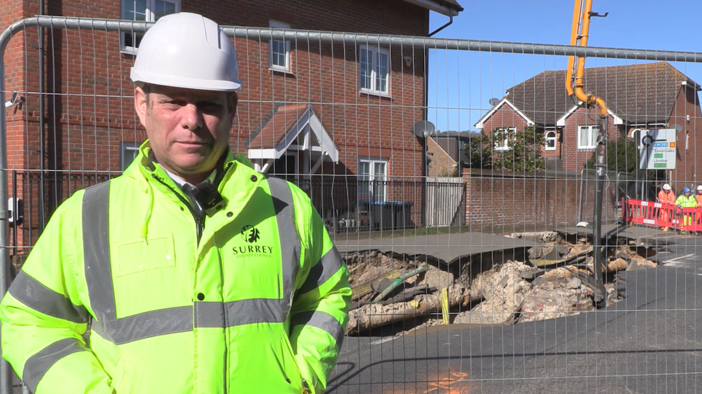 A man in a high-vis yellow coat with "Surrey County Council" written on the chest with the authority's emblem. He is wearing a white hard hat and standing in front of a metal fence with a large hole the other side.