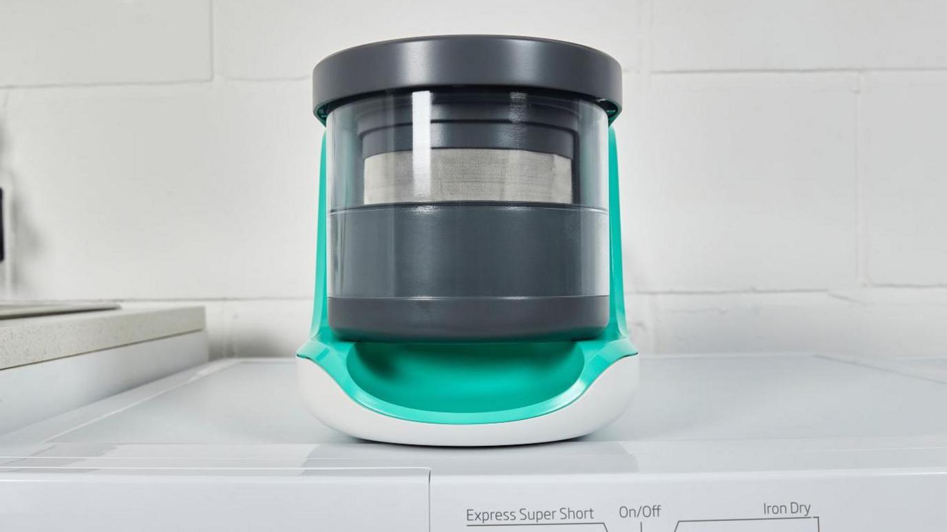 One of the filters made by Adam Root's company is placed on top of a white washing machine. It is cylindrical and white, turquoise and black in colour.