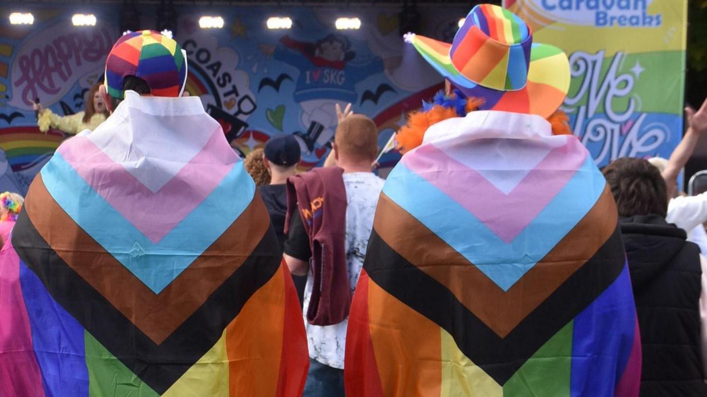 Two attendees are draped in identical Pride flags and stand with their backs to the camera.  Both wear different rainbow hats and are watching a performance on the main stage.