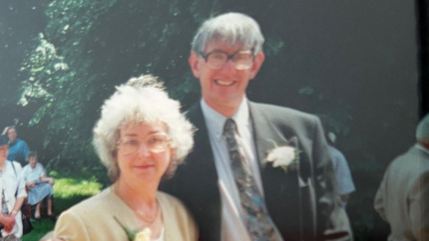 An old colour photo of Jean Turnbull and Raymond Turnbull, taken outside at a wedding or similar event. They both have large glasses and flowers in their button holes. Jean has white-grey hair to just above the shoulder and is wearing a yellowish tan light jacket. Raymond has grey hair brushed forward and wears a dark grey suit jacket over a very pale blue shirt and patterned tie. It is a sunny day and a large tree thick with leaves forms the background. A few other guests can be seen milling around.