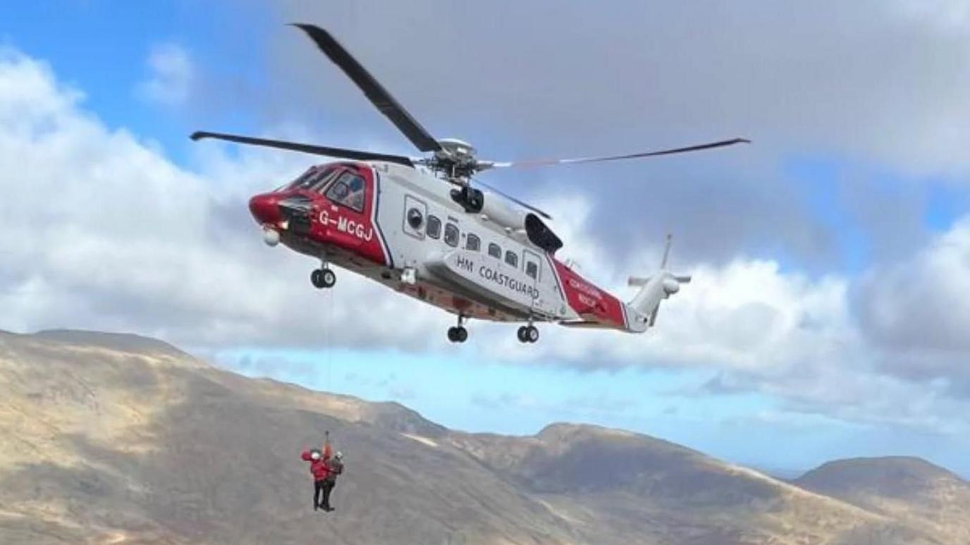 A red and white helicopter hovers in the mountains in north Wales, with people dangling on a winch line below it