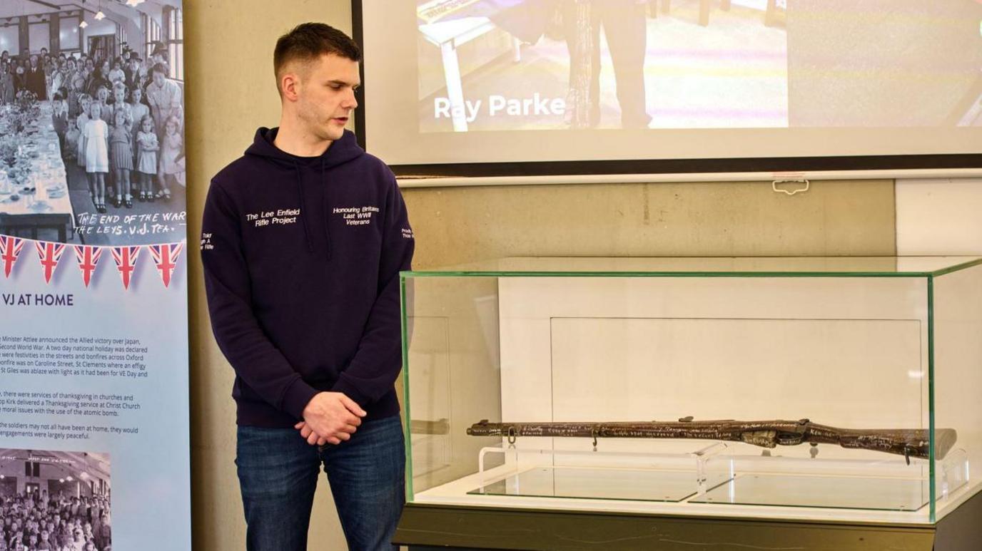 A man with a top branded Lee Enfield Rifle Project giving a presentation on the project. He is standing next to a glass display of a rifle with signatures of more than 130 World War Two veterans from across the forces.