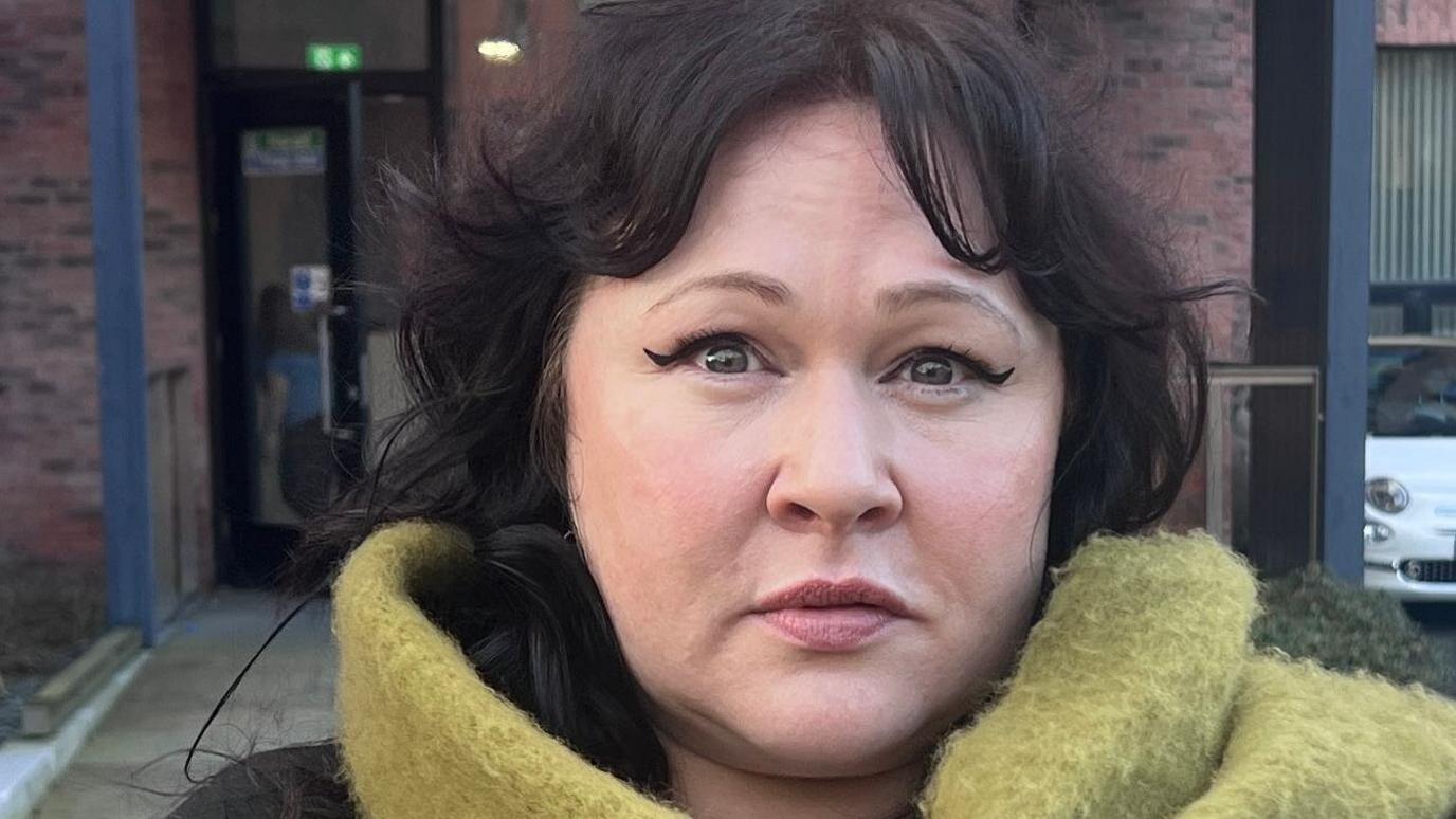 Sara Morrison stands outside the entrance to a red brick municipal building. Her shoulder-length dark hair is parted in the middle and her expression is neutral. She is wearing a black long-sleeved top and her green woollen scarf is around her neck and over her shoulder.