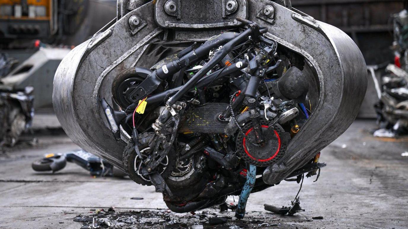 A large grabber of a scrapyard handling machine with several crushed scooters in its claws