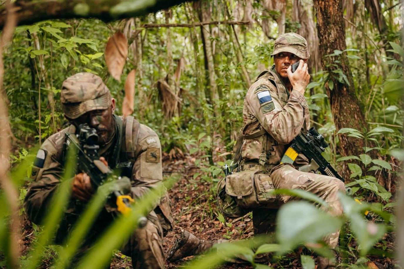 Two army soldiers armed with machine guns and dressed in camouflage crouching in the jungle while one tests a new smartphone.
