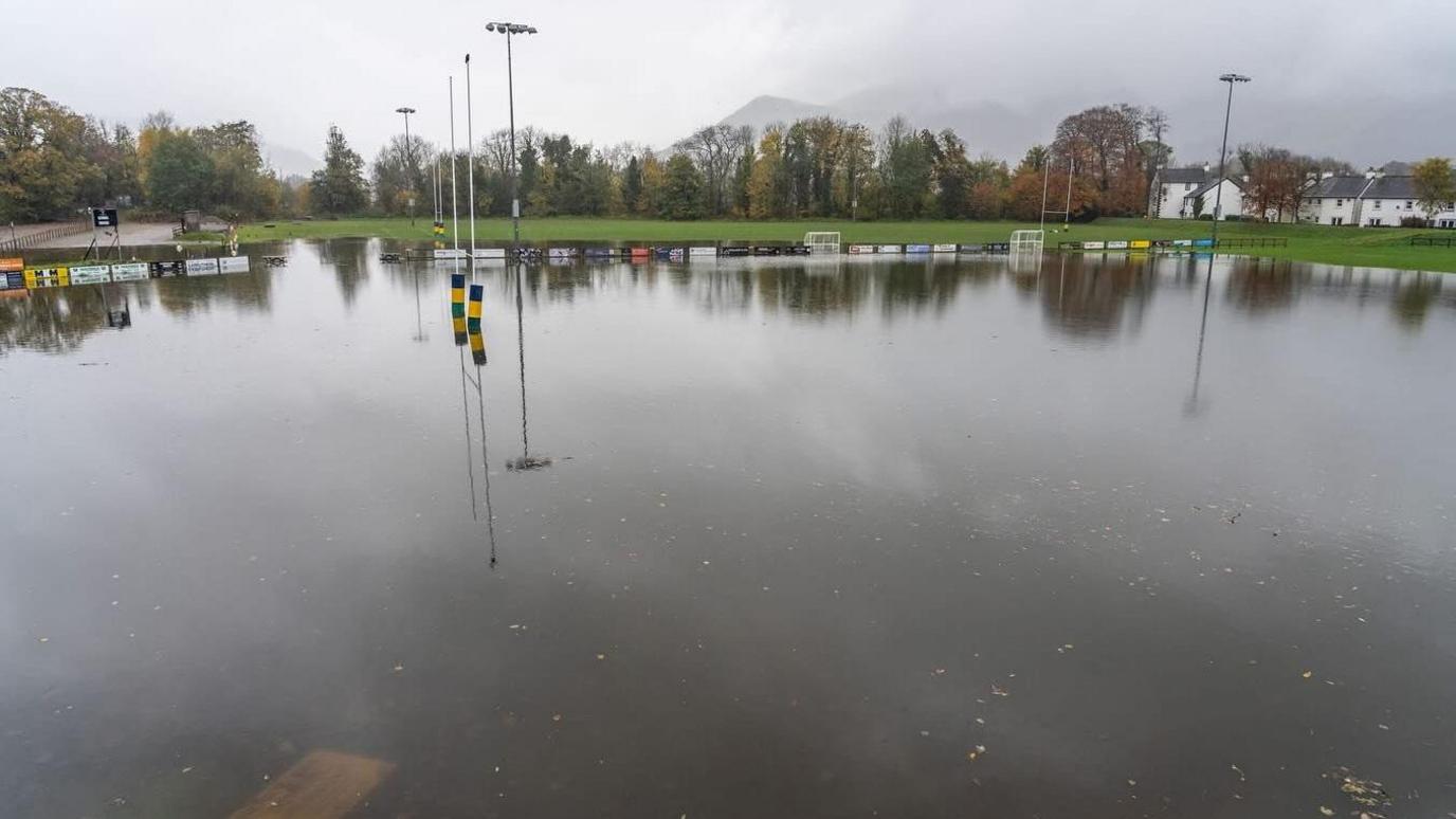 The pitch at Keswick Rugby Club is completely submerged by flood water, which is very deep. There are lots of rain clouds in the sky with mist appearing from the fells.