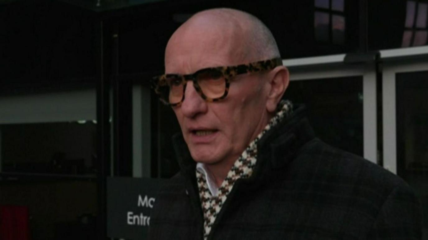 Colin Neill wears a black checked jacket and checked scarf. He has grey hair and glasses.