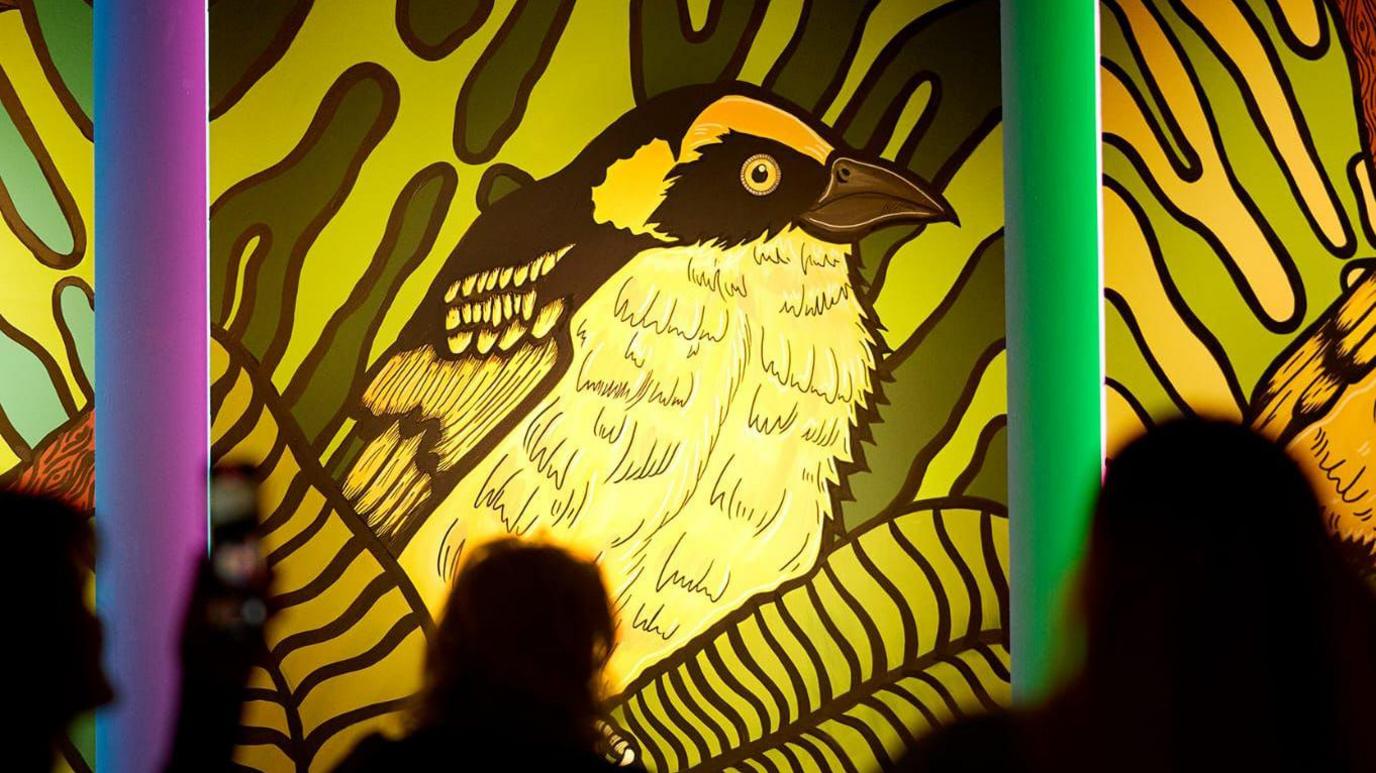 An art work of a bird was on a wall with a light shining behind it. A purple and green poles were standing either side and people were stood looking at the piece of art.