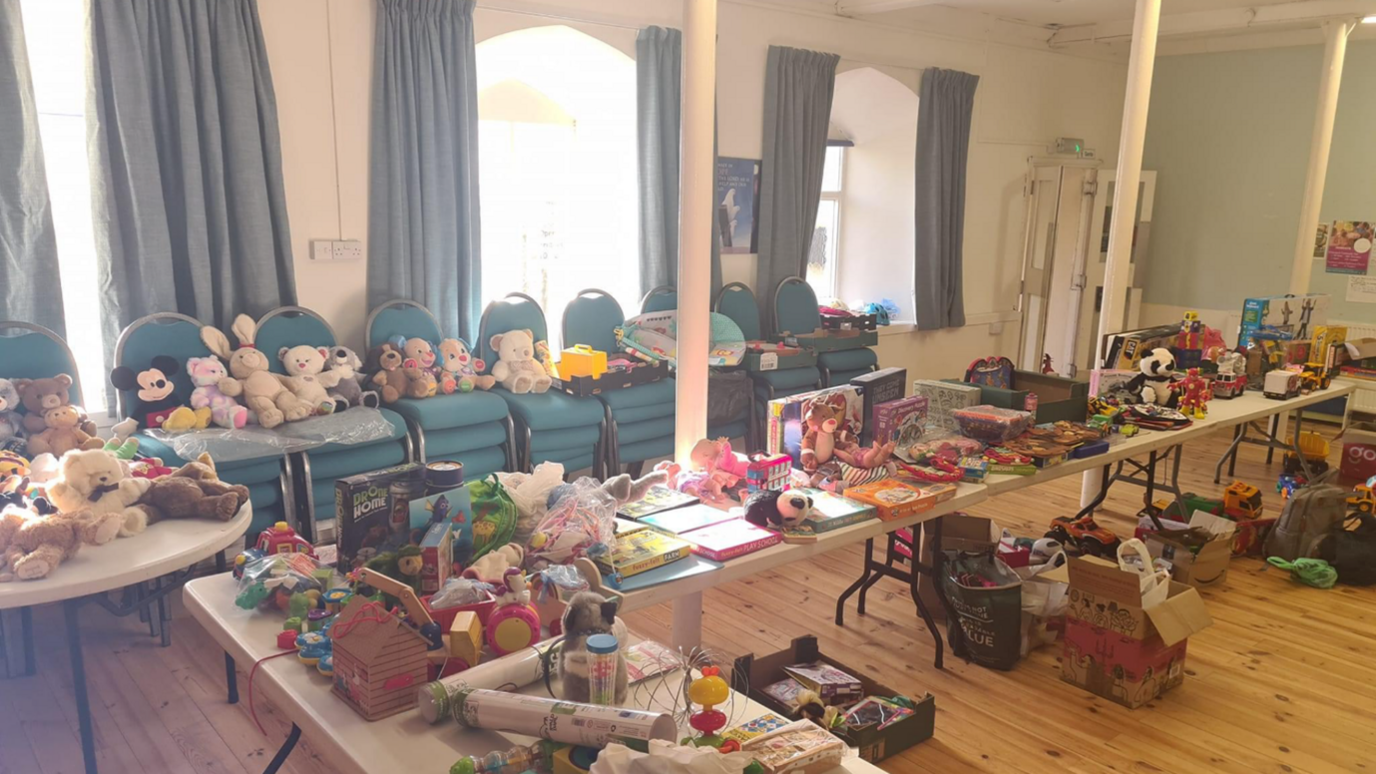 Tables filled with toys and teddies at the church. There are teddies displayed on stacked blue chairs. The floor is brown laminate and there are three large windows with curtains behind the chairs