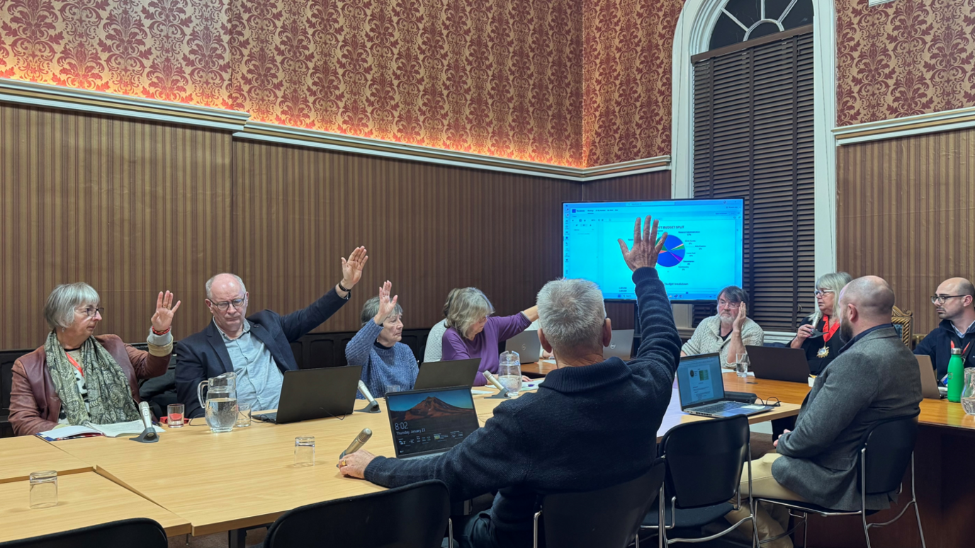 A group of councillors in a town hall room sitting round a table in front of a screen with a power point on it. Some councillors have their hands raised