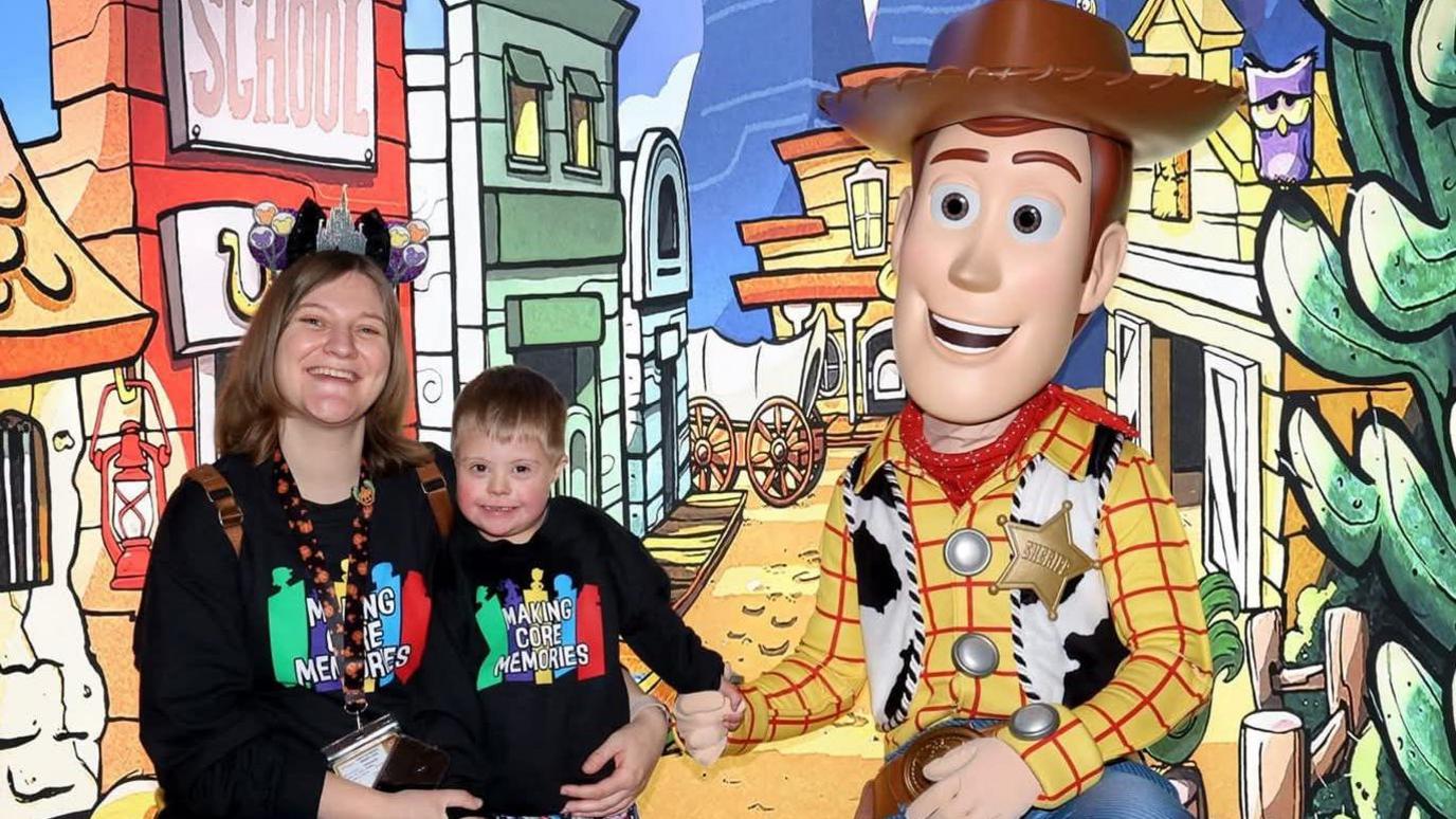 Jaxson's mum has shoulder length brown hair and is wearing a pair of black mickey mouse ears as a headband. She is wearing a black jumper that reads "making core memories", and has her arm around her son, a young boy with light brown hair. Jaxson is holding hands with a person dressed as Woody from Toy Story - who is a cowboy with red and yellow top and a gold star badge
