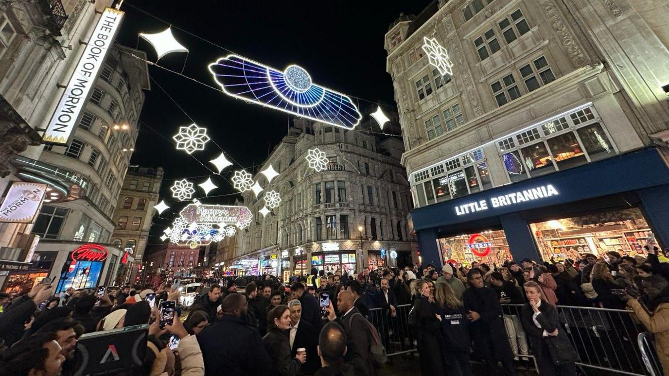 A large crowd gathered under the lights and the Mayor of London can be seen having his photo taken under the lights. The theatre with the Book of Mormon is to the left and a shop called Little Britannia to the right. 