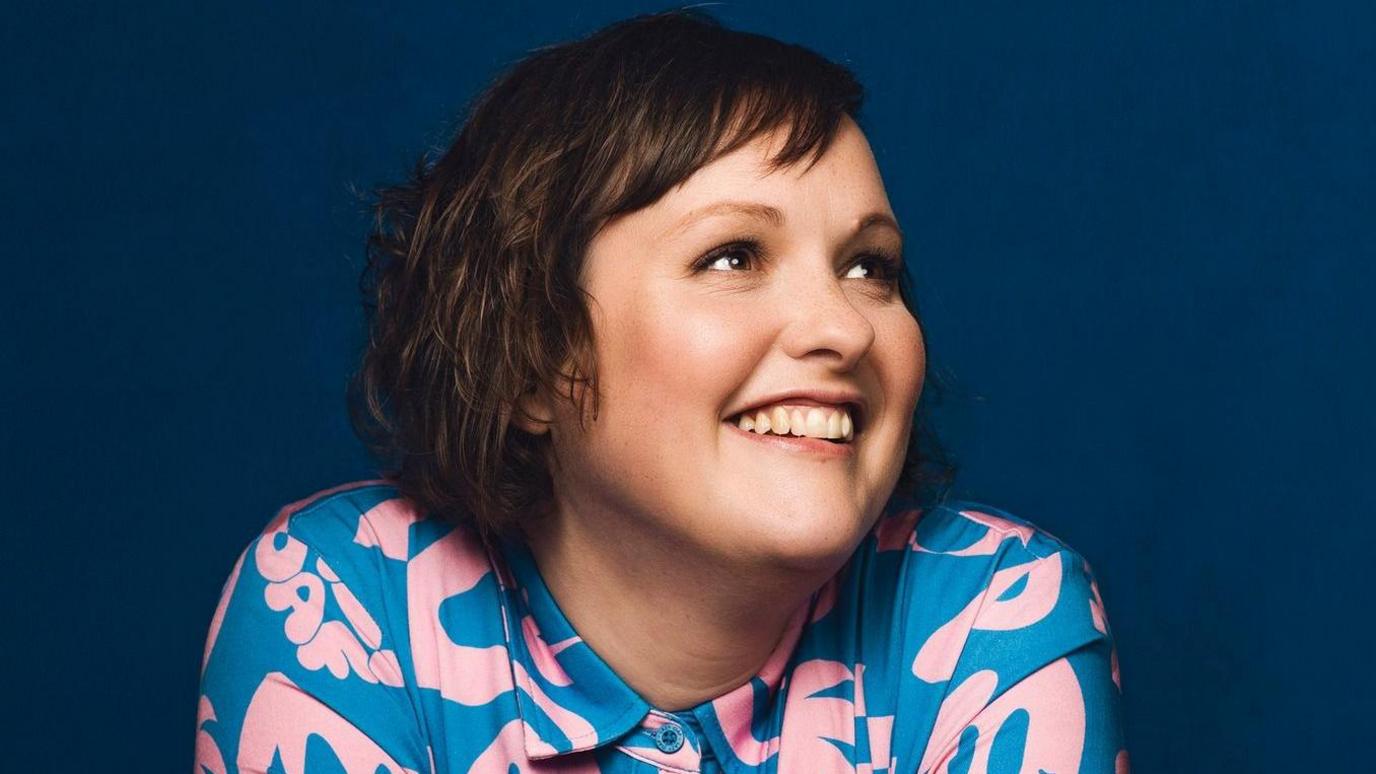 Josie Long smiling with short brown hair and a pink and blue top