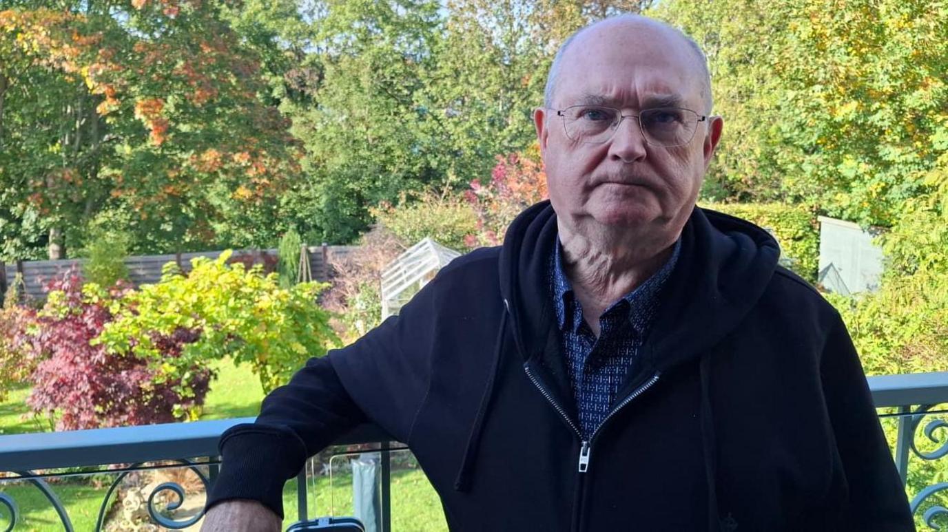 A man looks into the camera with a neutral expression on his face. He is standing on a balcony overlooking a garden with a woodland in the background.