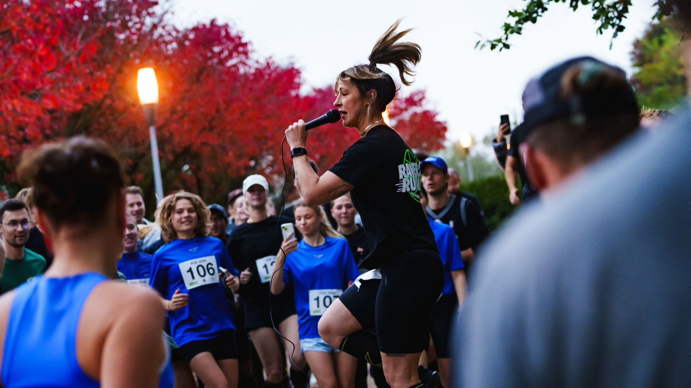 Jo, wearing black running gear and standing with a microphone in her hand looking at a group of runners with race numbers bibs on, getting them warmed up before a run