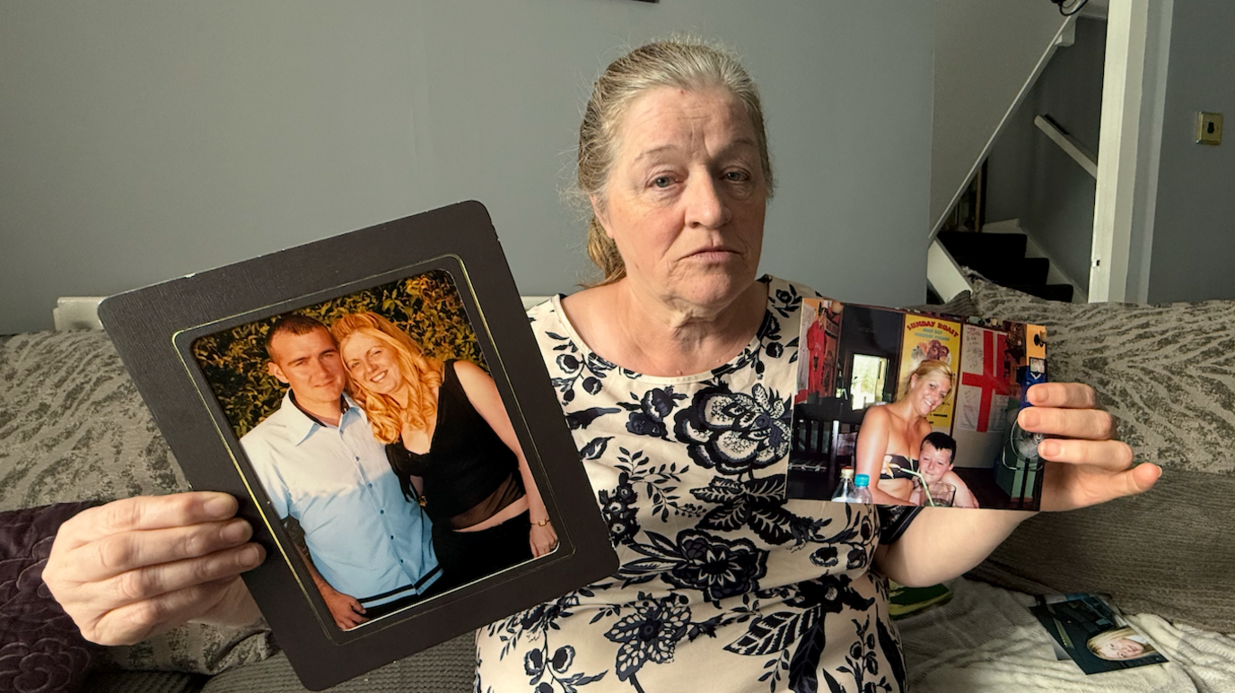 Margaret Bramall wearing a patterned dress holds up family photographs. The one in her right hand shows her daughter Lynsey and her partner, while she holds a photograph in her left hand showing her daughter smiling with her young son.