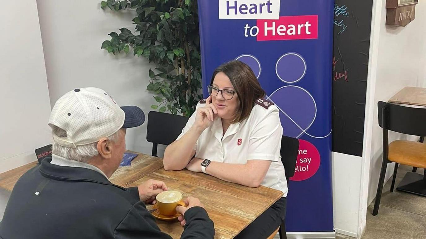 Major Alison Friday dressed in Salvation Army uniform sits at table in The Canterbury Bakery talking to an elderly man over a coffee