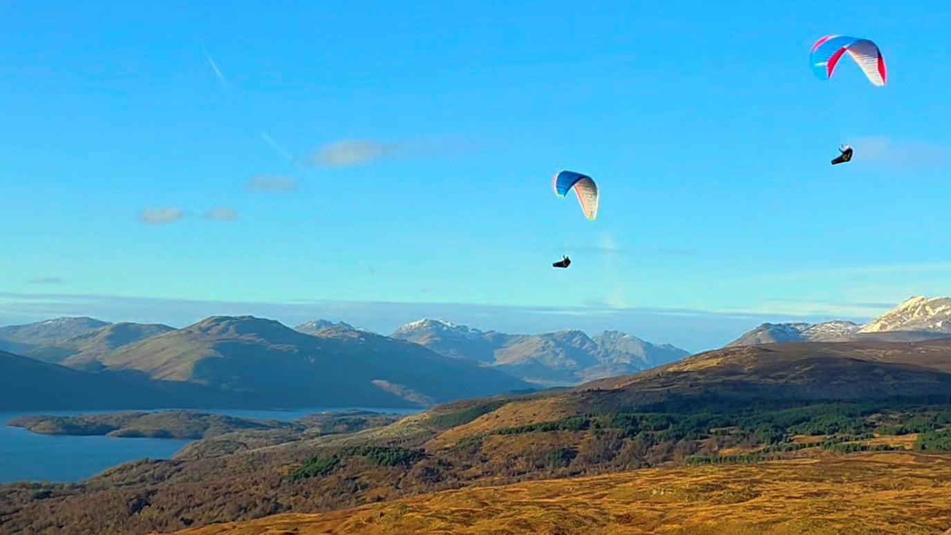 Three colorful paragliders soar above rolling hills and a distant snow-capped peak.