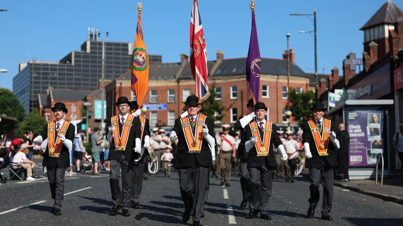 Five men wearing Orange Order sashes are leading a march the middle of a road.  Three behind them are holding flags. Crowds are in the background. 