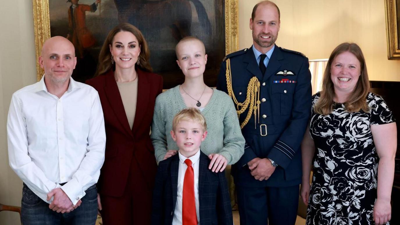 Liz Hatton, pictured with the Prince and Princess of Wales. The Princess, who is smiling at the camera, wears a burgundy blazer and beige top. She stands next to Ms Hatton, who is wearing a light green jumper and necklace. Her hands rest on the shoulders of a young, blond boy in front of her, who is wearing a dark suit and red tie.