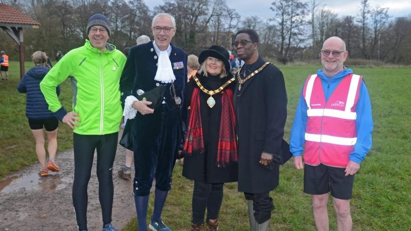 Five people are stood in a line looking at the camera. On the left is a man in a yellow running coat, hat and black running leggings. Next to him on his right is a man in a ceremonial blue jacket with a white cravat, blue tights and trainers. On his right is the mayor and she's wearing her ceremonial necklace, with a black hat, long black coat and black jeans. On her right is the deputy mayor in his ceremonial chains, a long black coat and wellington boots. Jamie Hill is on the right, and wearing a blue jumper with a pink high-vis over it, as well as black shorts.