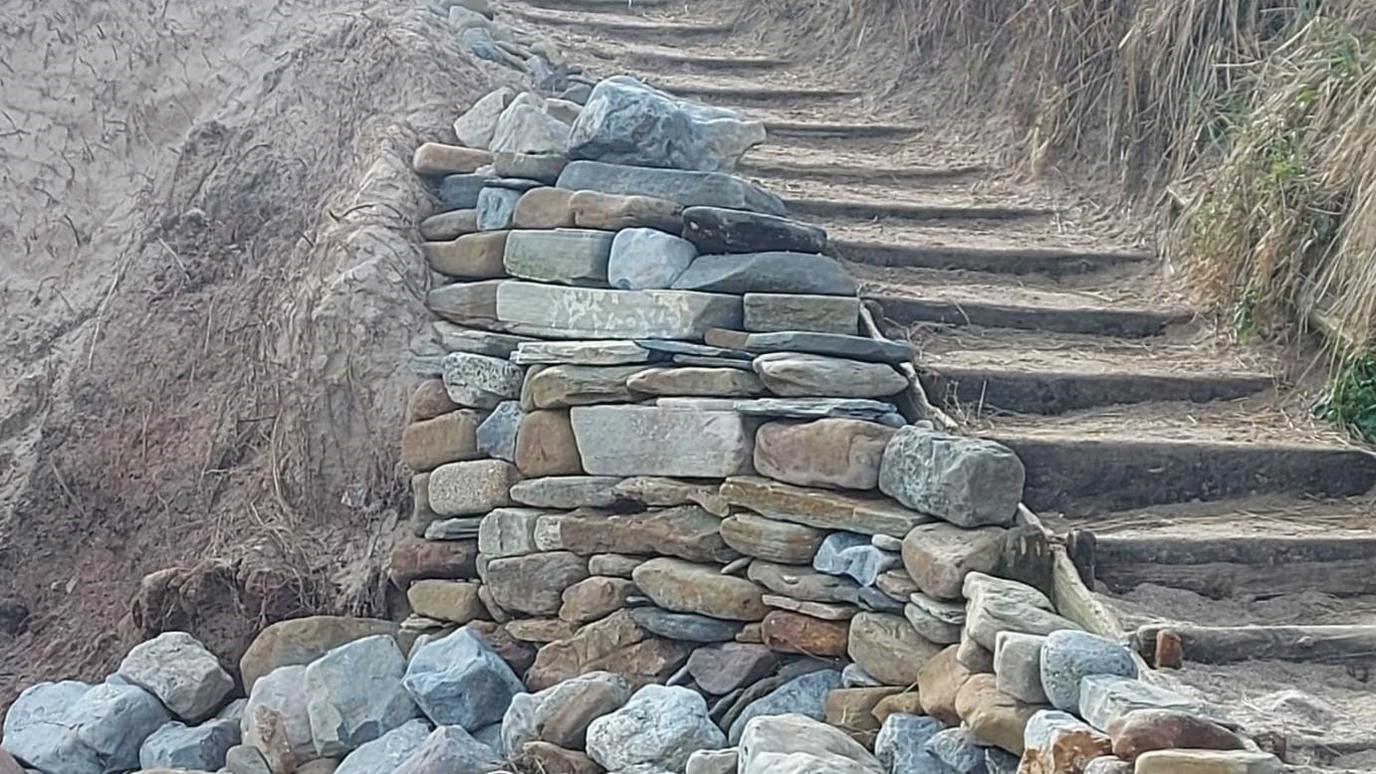 The picture shows a half constructed wall with a sandy bank beyond and a series of steps up to the bank 