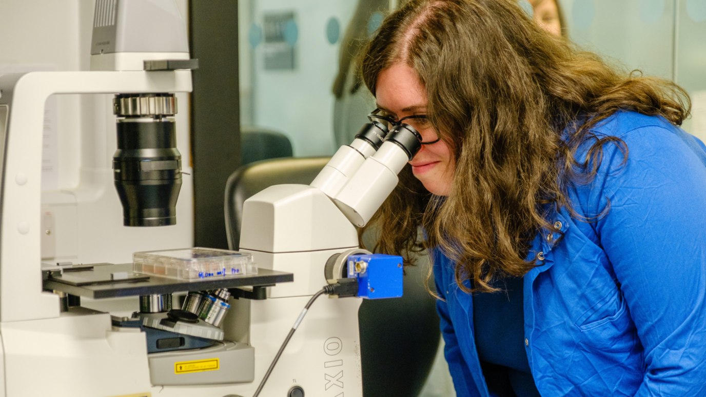 Dr Osborn, who has long dark hair and is wearing a bright blue shirt,  is looking down a microscope in a lab