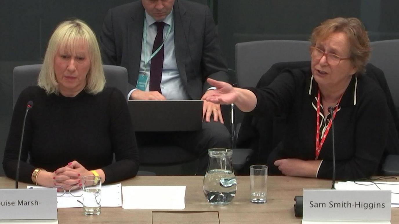 Dwy ddynes mewn dillad du yn eistedd tu ol i fwrdd yn un o bwyllgorau Senedd Cymru, gyda'u henwau ar bapur o'u blaenau - Anna Louise Marsh-Rees (chwith) a Sam Smith-Higgins (dde)