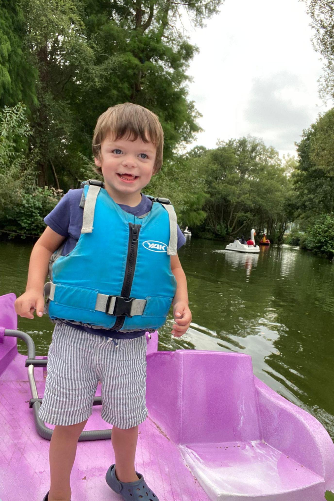 A little boy with light brown short hair is smiling while standing on a pink pedal boat on a lake. He is wearing a blue T-shirt, striped blue shorts and a turquoise life jacket. There are large pedal boats shaped like swans and other birds behind him.
