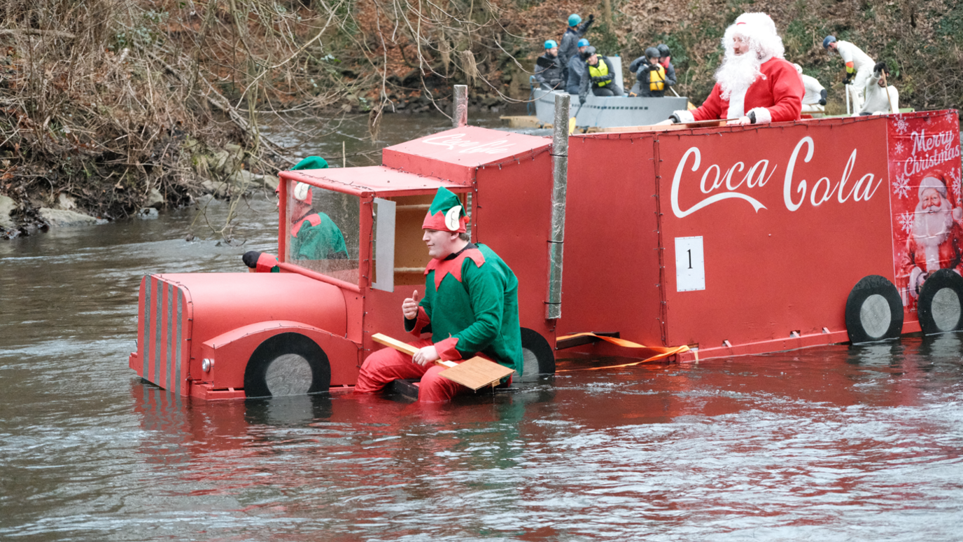 Two elves with oars sit at the front of a raft shaped like a red Coca-Cola lorry. A person dressed as Santa Claus sits at the top.