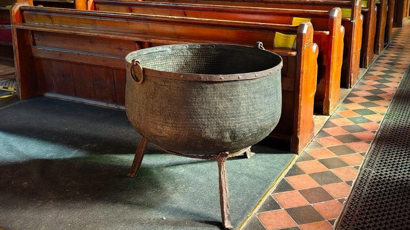 A large black copper cauldron on a rusty metal stand with three legs. It is in the nave of a church. In the background are rows of pews