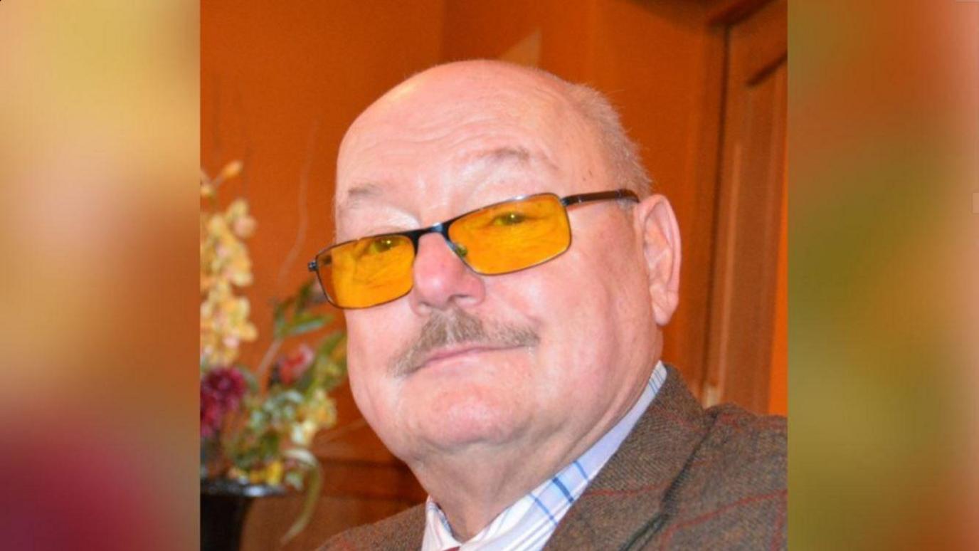 Henry Thirsk, smiling into the camera wearing a tweed jacket and a white and blue shirt. He has a moustache and his wearing yellow-tinted glasses. 