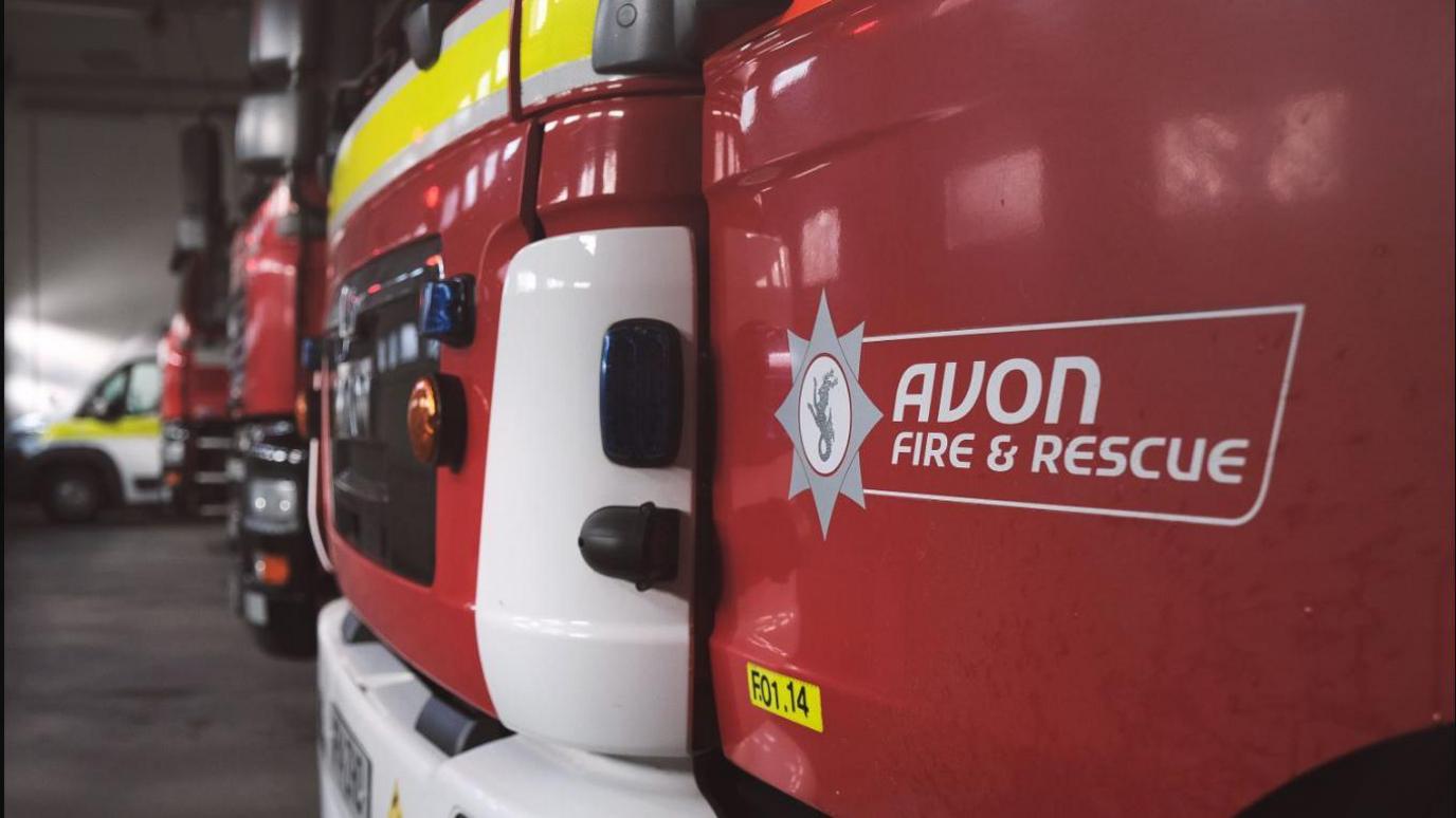 Photo showing the front bumper and bonnets of a line of fire engines, lined up inside a station. The one nearest to the camera has "Avon Fire and Rescue" in white writing on a red panel