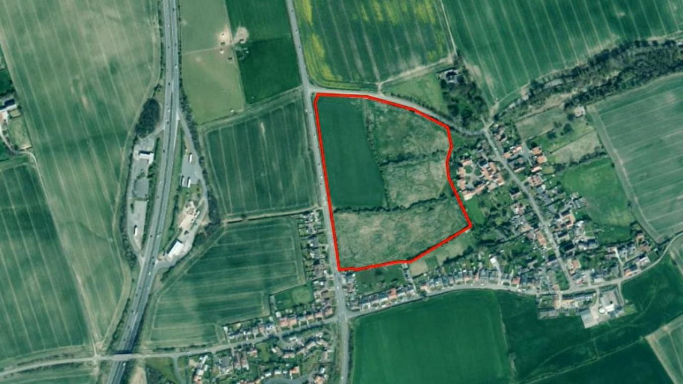 An aerial map of the proposed site highlighted by a red boundary. The boundary is surrounded by green fields with a small settlement to the right. To the left there is a major road.