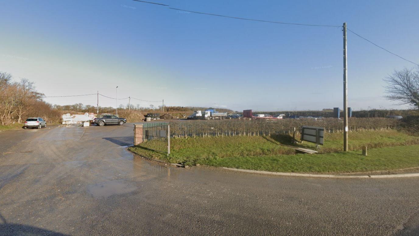 A driveway leading up to a car park area shown on the left of the photo. A few cars can be seen in the car park in the distance. Green railings can be seen on the right of the driveway with green grass in front of it. 