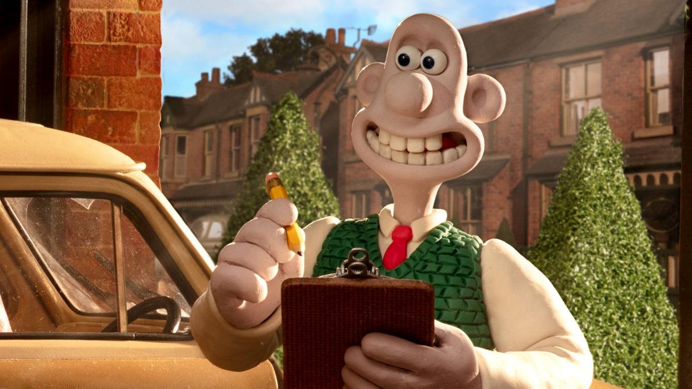 The animated character Wallace from Wallace and Gromit smiles and looks away while holding a clipboard and pencil, with the front of a yellow car and a row of houses on a street with Christmas trees in the background, in a still from the upcoming Wallace and Gromit feature film 'Wallace and Gromit: Vengeance Most Fowl'