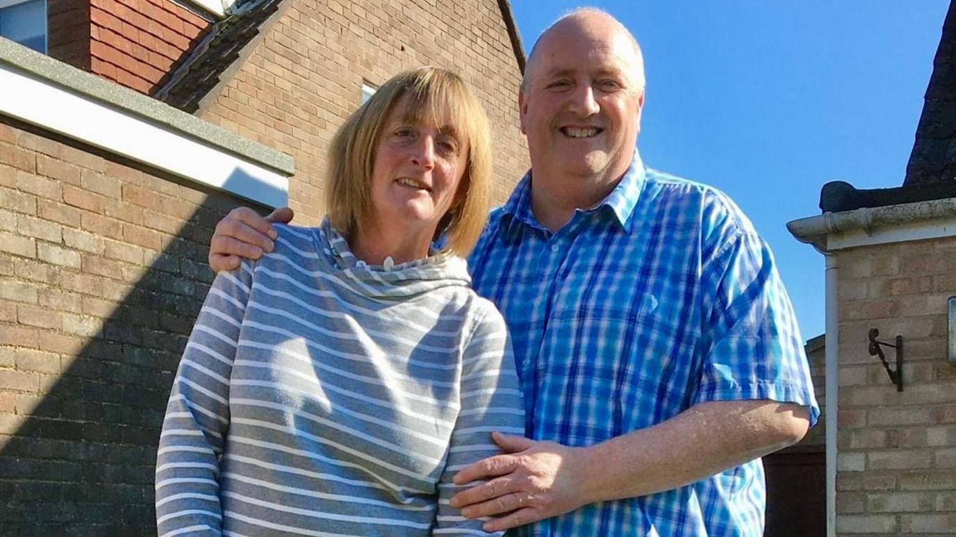 Photo of Lisa and Spencer. They are pictured outdoors, standing next to each other. Lisa has short blonde hair and is wearing a grey and white stripe jumper. Spencer is standing next to her with one arm around her, and the other touching her arm. Spencer is bald and is wearing a blue check shirt. They are both looking at the camera and smiling