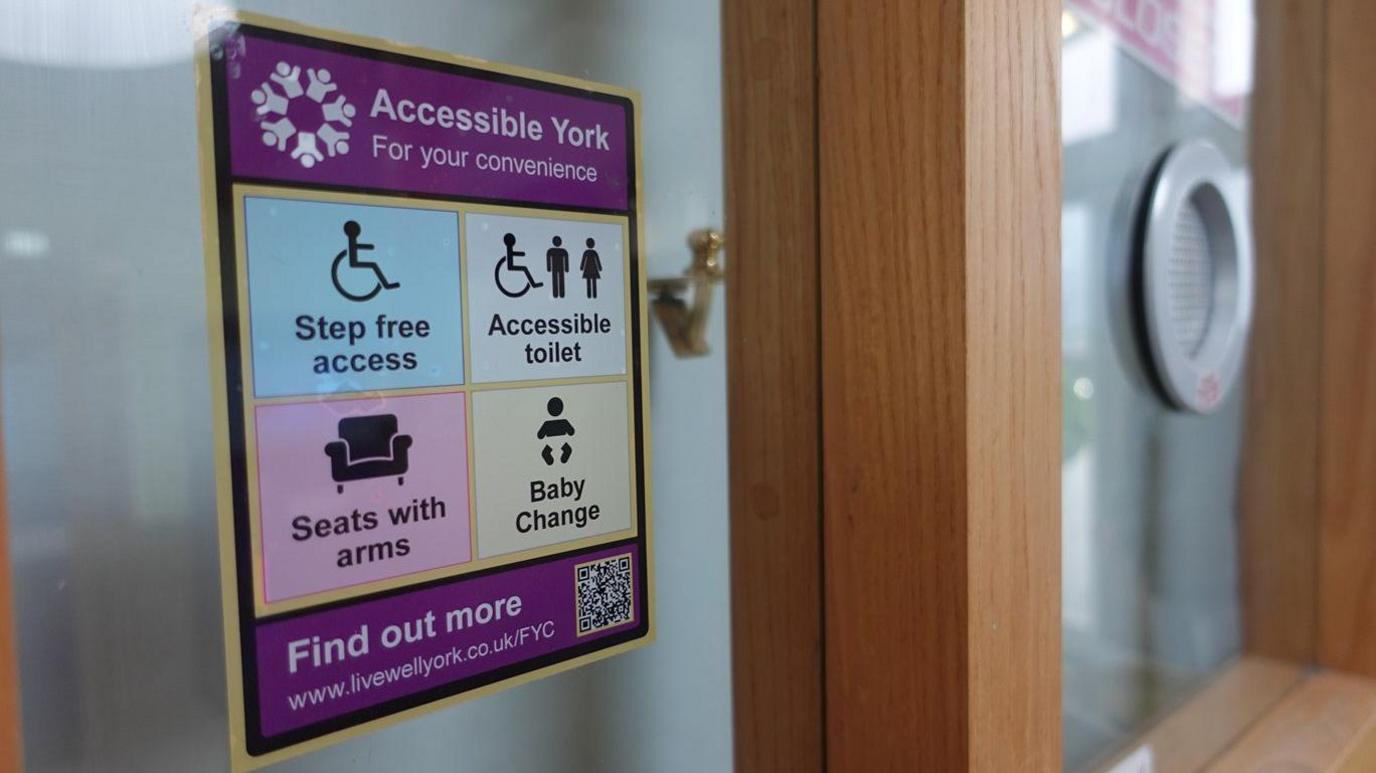 An Accessible York sticker on a frosted glass door. The sticker is rectangular, split into four sections with symbols and writing indicating the premises have step-free access, an accessible toilet, seats with arms and baby changing facilities.