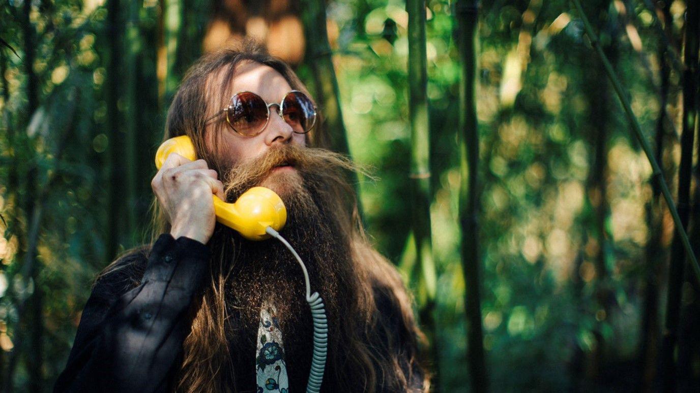 Richey Powers has long hair, a long moustache and a long beard. He is wearing round sunglasses and speaking into a yellow telephone. He is wearing a black jacket, black shirt and blue flowery tie.