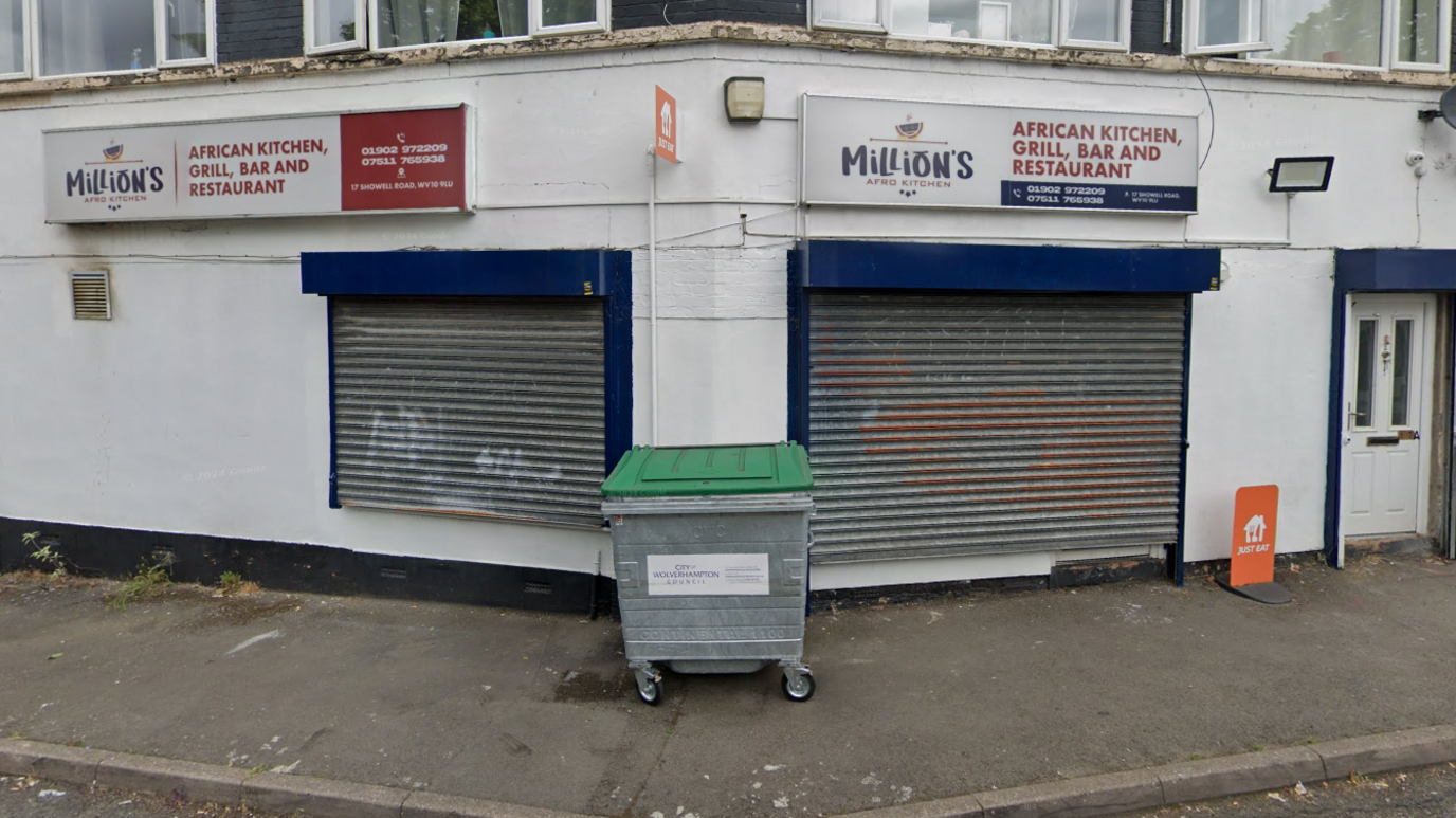 The business is pictured during the day with metal shutters pulled down. The building is white and with blue paint above the shutters with a white sign.