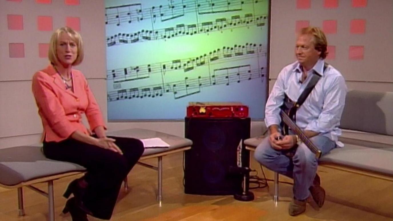 An archive image of Sally Taylor and Mark King. They are both sitting on grey stools and King is holding a guitar