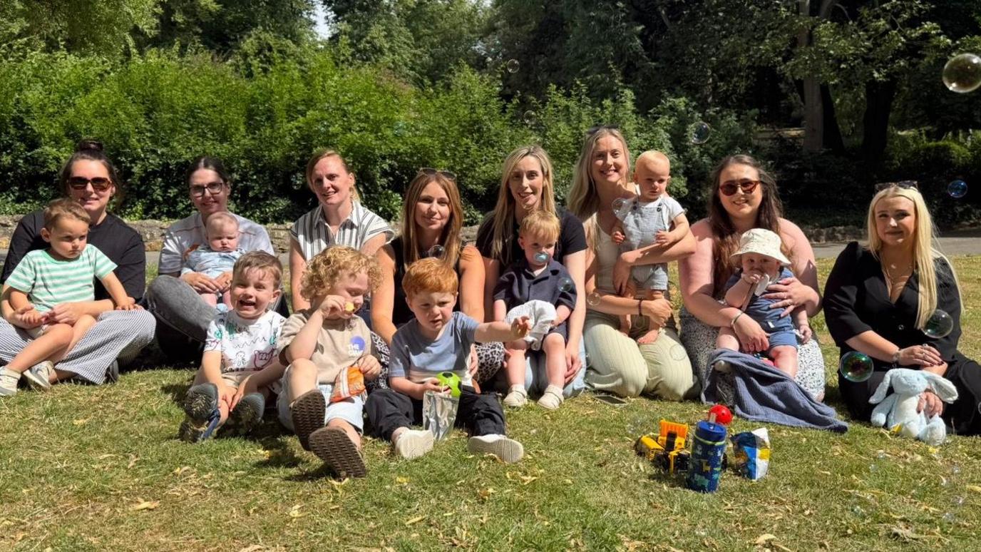 Eight women sitting in a park and smiling at the camera on a sunny day. In front of them are eight boys who are sitting down on the grass or on their mum's knee. Some are eating, others are looking at a bubble machine in the distance. Children's toys can be seen.