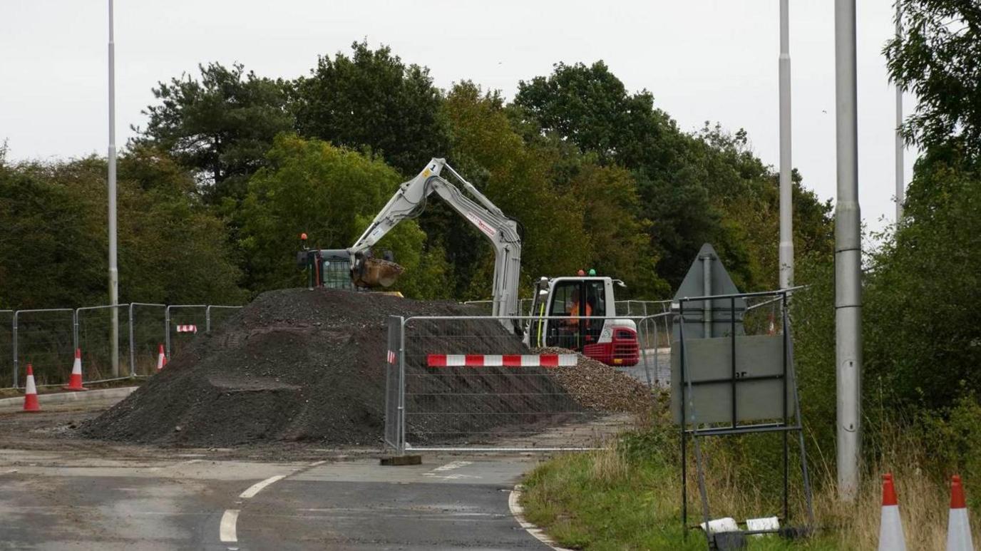 A large pile of an unspecified material (soil or sand) takes up the centre of the photo and is within a sealed off section of road. Two diggers are pictured working with the material