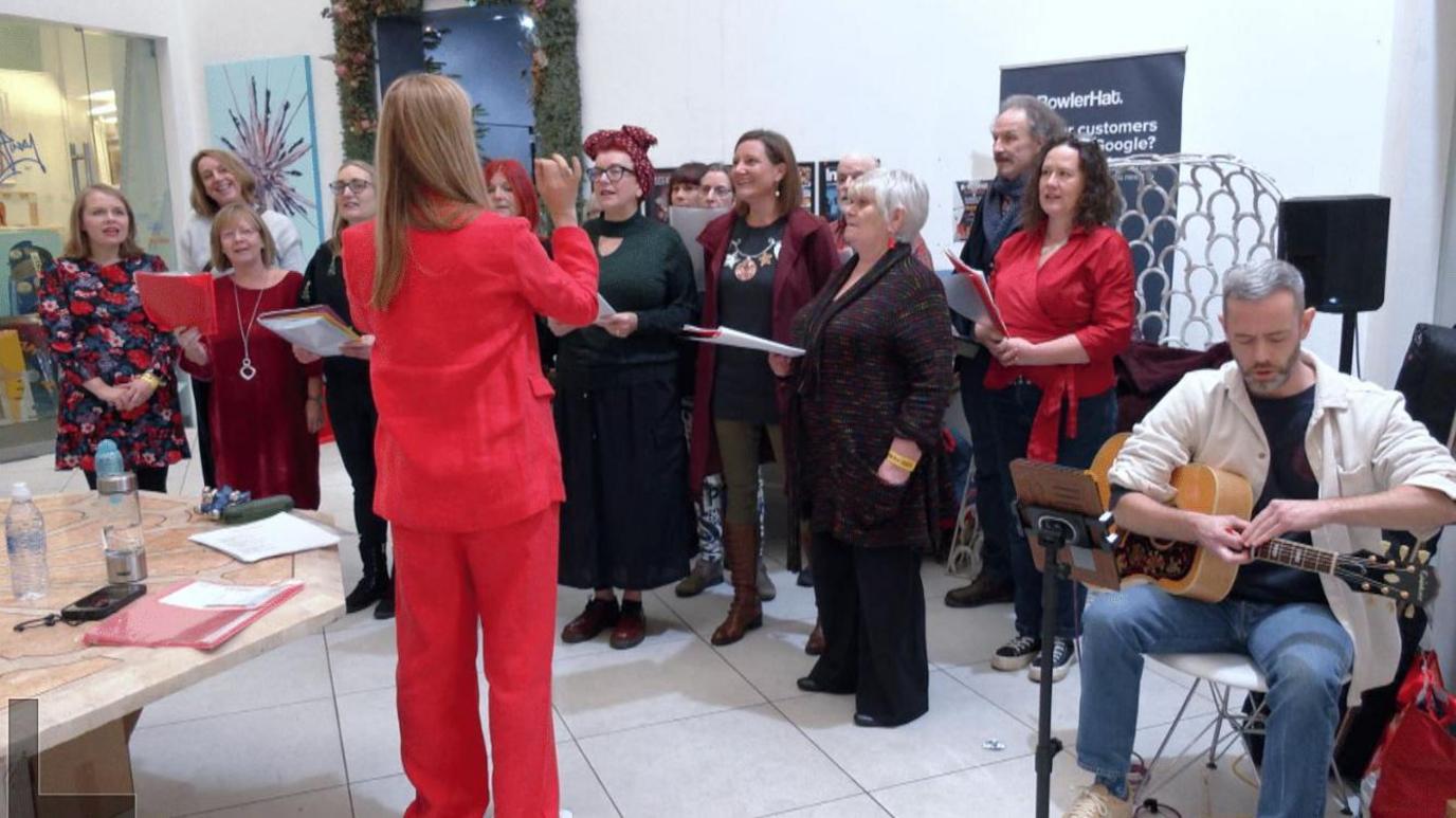 12 singers stood in two rows, looking at a woman in a red suit whose back is to the camera and her arms are raised as she conducts. A man sits on the right playing a guitar. The choir is in a building with white tiled floor and white walls with artwork.