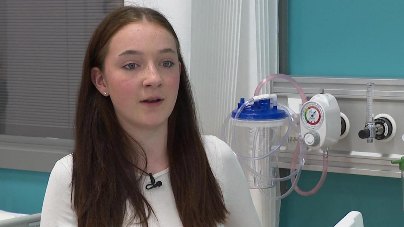 Miah, with long brown hair and wearing a white top, wants to be a children's nurse. She speaks in front of equipment designed to simulate a hospital bed