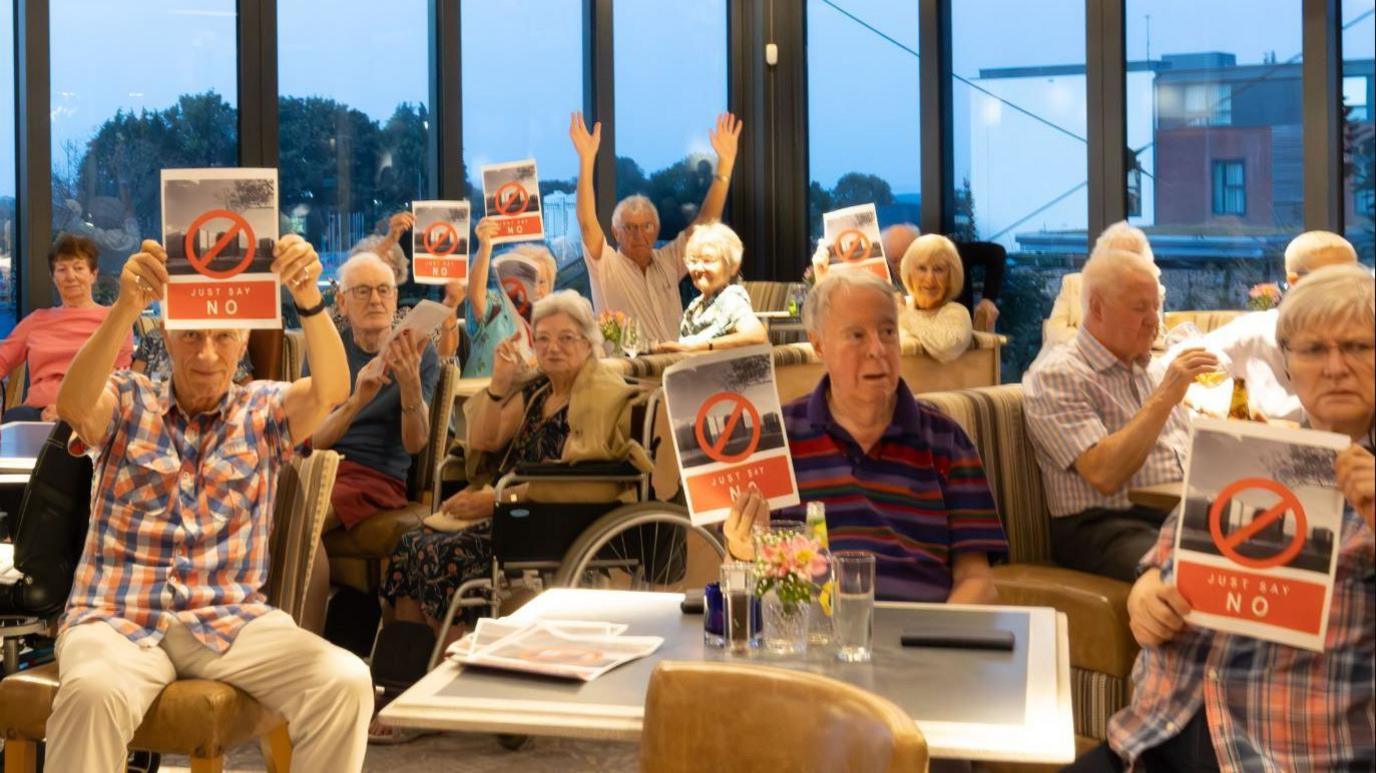 A group of retirement village residents are sitting around tables in their building. Some of them are holding signs which say ' Just Say No' 