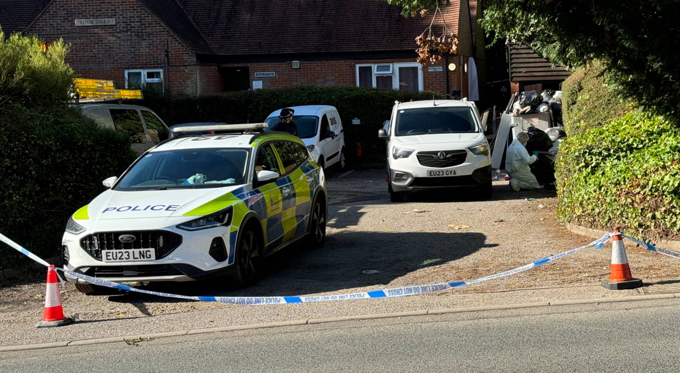 A police car and forensics vans parked on a driveway. Police tape blocks entrance to the property. 