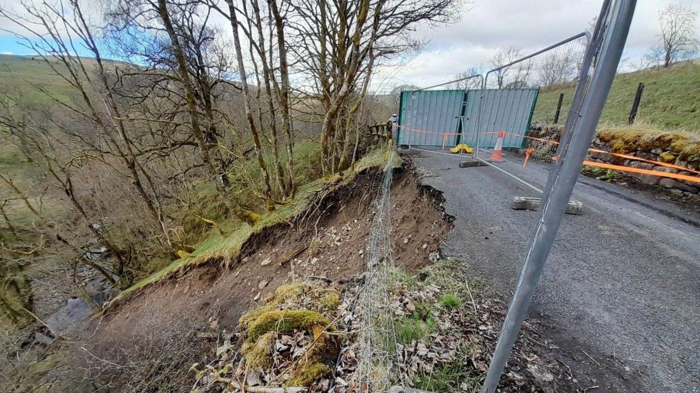 A single-track rural road is damaged on one side. A previous landslide has exposed mud on the hillside leading down to a body of water at the bottom of a valley. Metal fences and bright orange tape has been placed around the affected area.