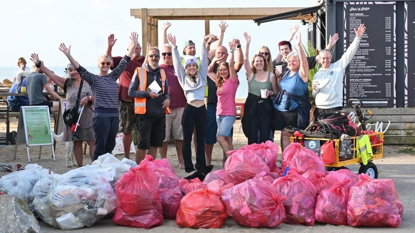 A group of about 15 people stood in two rows with their arms up in the air. Behind them is the beachfront and picnic benches, where some more people are sat. In front of them are piles of full rubbish bags and a yellow trolley filled with equipment such as high viz vests and litter pickers.