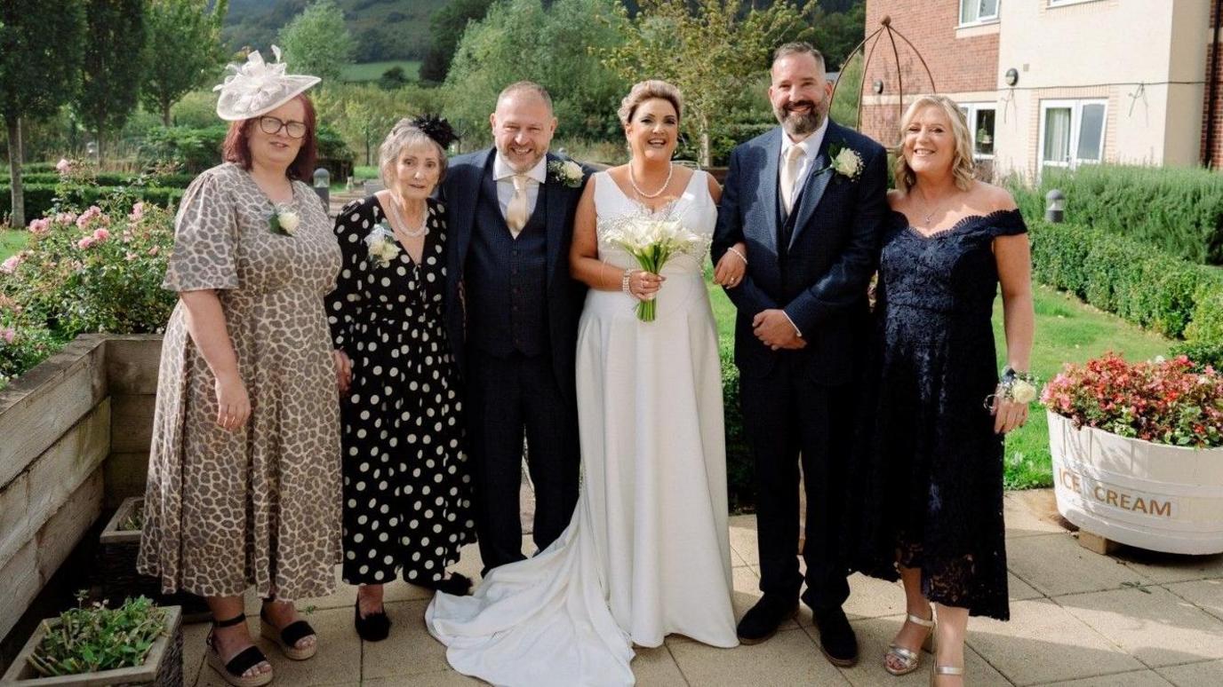 The couple and their family, including Rachel's mother, Ruth, smiling outside in the garden of the carehome. The woman far left is wearing a leopard print dress with a large white hat, she has dark reddy-brown hair. Next to her is an older woman in a black and white dress. To the right of her is a balding man with a blue three piece suit and cream tie and flower. To the right of him is a woman in a white wedding dress holding a bunch of white flowers. Next to him is a man who has short grey-black hair and a blue three piece dark blue suit, he also has a cream tie and flower. And finally to his right is a woman with shoulder-length blonde hair and a blue off the shoulder flowery dress. They are stood in the garden and you can see trees, grass and flowers behind them. To the right is a cream and right bricked building.